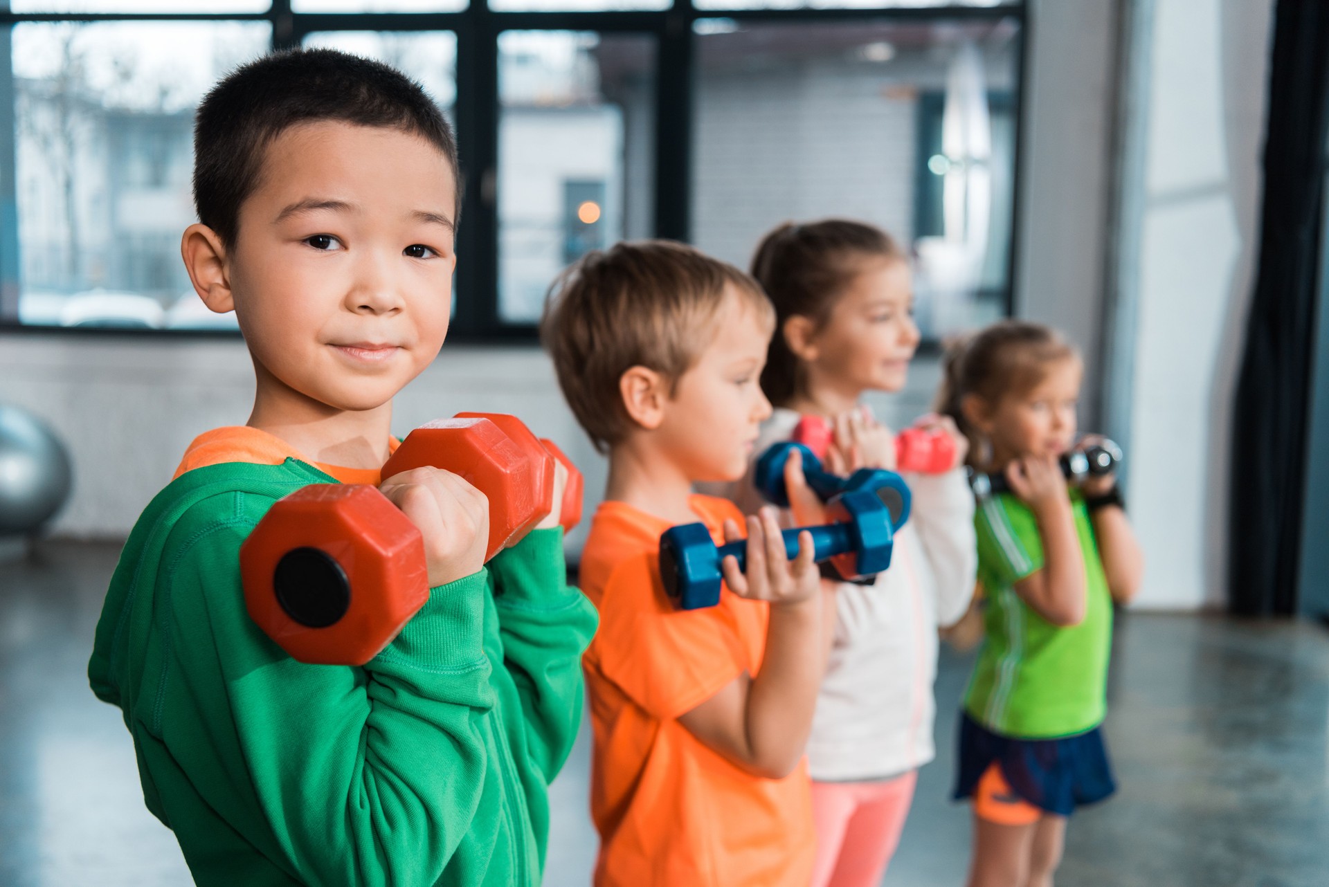 Selective focus of multiethnic children lined up, holding dumbbells in gym